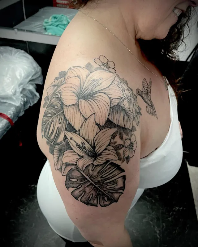 Side view of a shoulder tattoo with tropical flowers, leaves and a hummingbird.