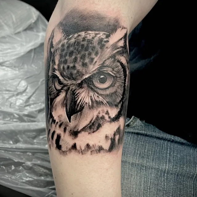 A shaded close-up owl head tattoo on a forearm.