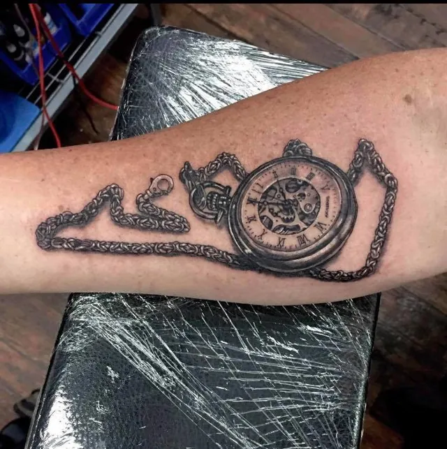 A shaded pocket watch and chain tattoo on a forearm.